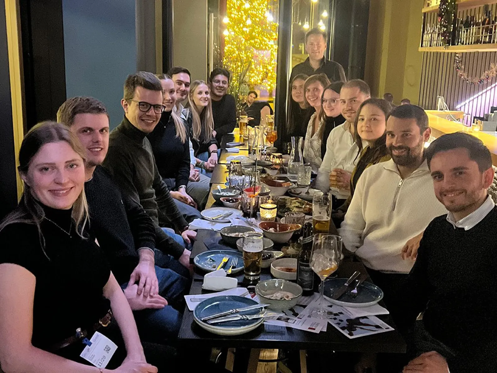 Eine Gruppe von Alumni der Universität Liechtenstein sitzt in einem Restaurant an einem langen Tisch, lächelnd und in geselliger Runde. Auf dem Tisch stehen Getränke und Teller mit Speisen. Die Atmosphäre ist warm und einladend.