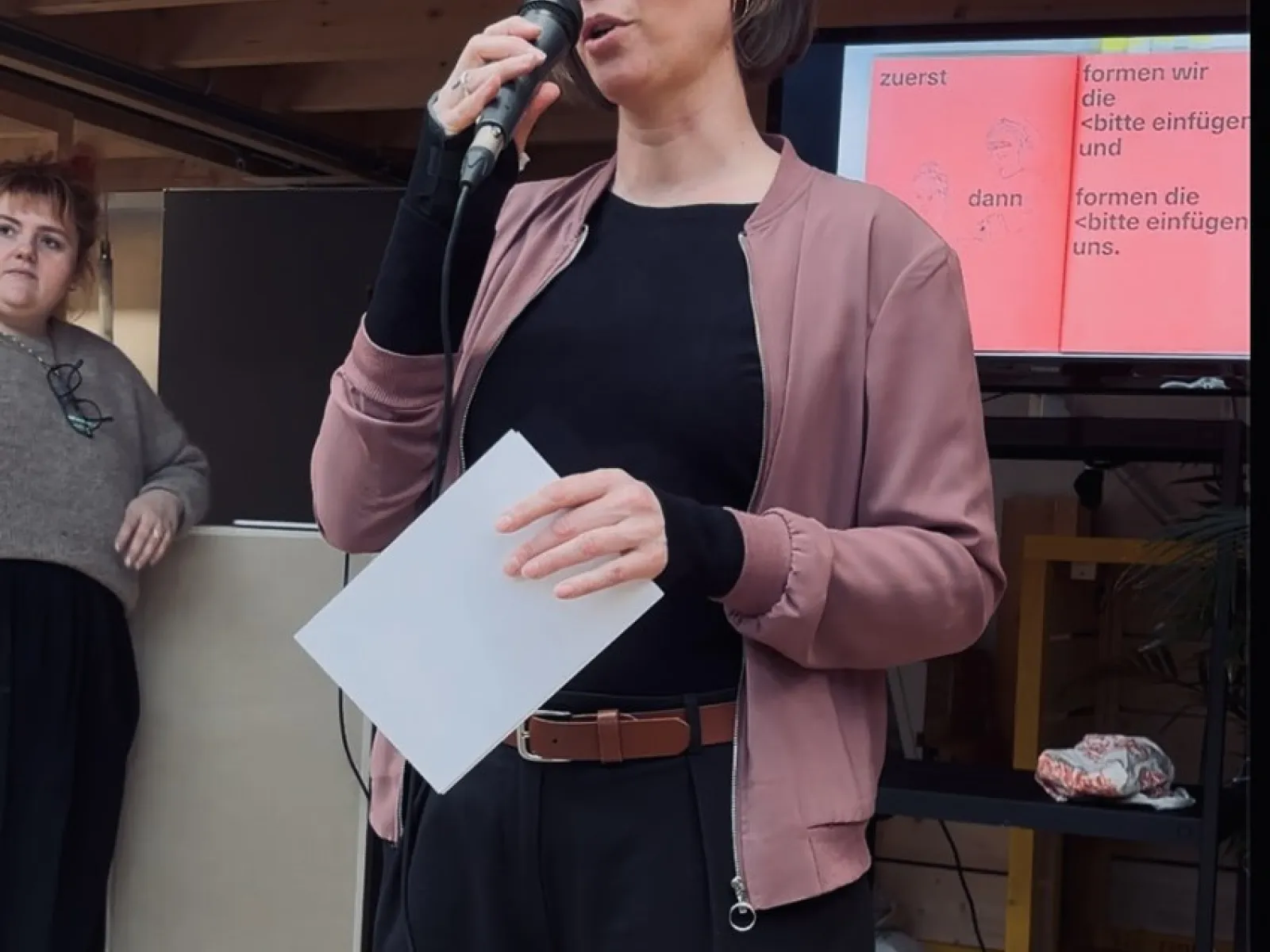 Eine Person hält ein Mikrofon und spricht vor einem Publikum. Sie trägt eine rosa Jacke und hält ein Blatt Papier. Im Hintergrund ist eine Präsentation auf einem Bildschirm zu sehen. Eine weitere Person steht links daneben und hört zu.