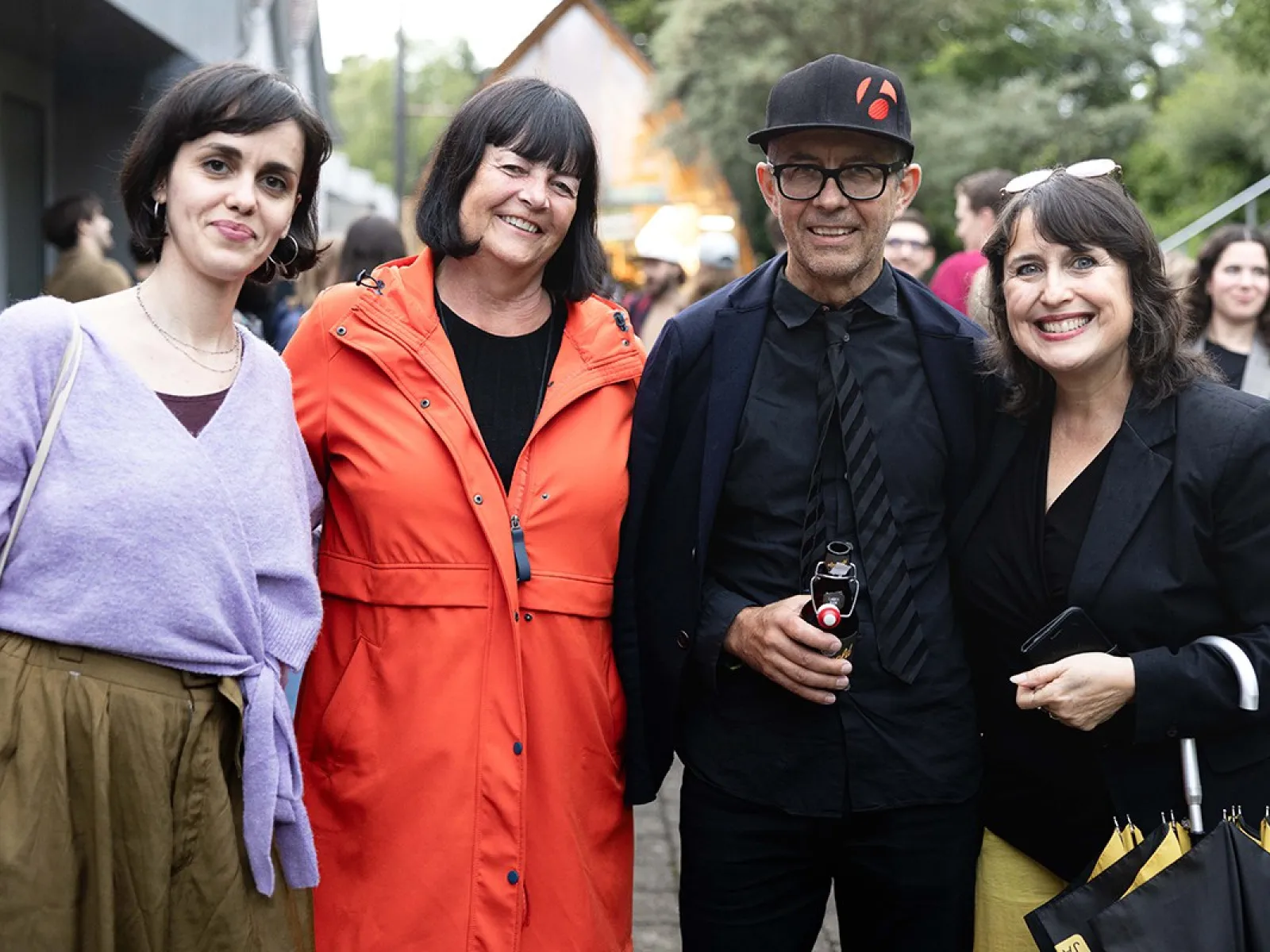 Vier Personen stehen lächelnd nebeneinander bei einer Veranstaltung im Freien. Sie tragen unterschiedliche, farbenfrohe Kleidung. Im Hintergrund sind weitere Menschen und Bäume zu sehen.