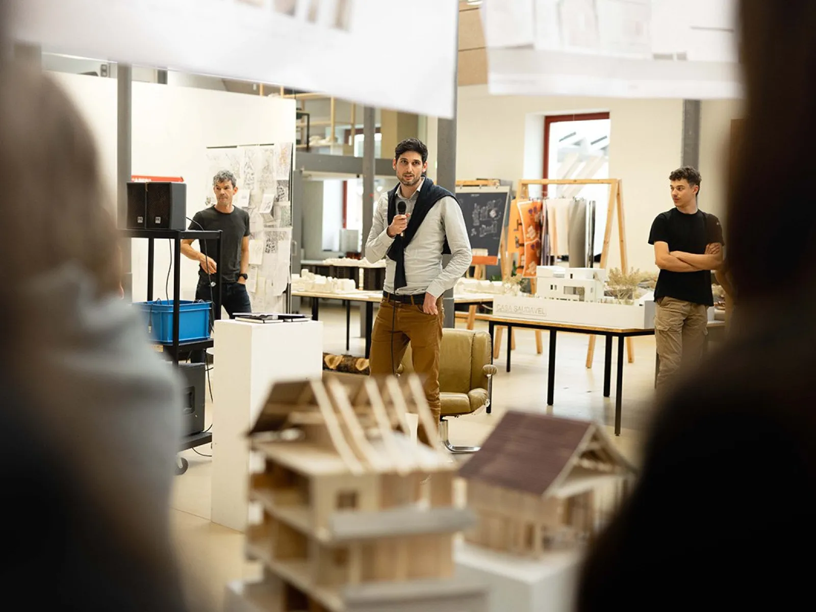 Ein Redner hält einen Vortrag in einem Ausstellungsraum der Universität Liechtenstein. Im Vordergrund sind Architekturmodelle zu sehen. Zuschauer hören aufmerksam zu.