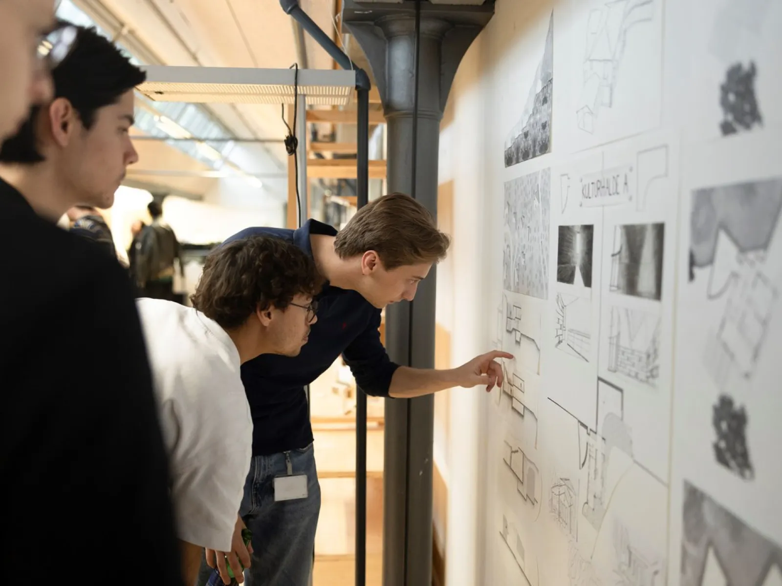 Drei Personen betrachten und diskutieren Architekturzeichnungen an einer Wand in einem modernen Innenraum. Eine Person zeigt auf Details der Zeichnungen. Die Szene vermittelt eine Atmosphäre des Lernens und der Zusammenarbeit.
