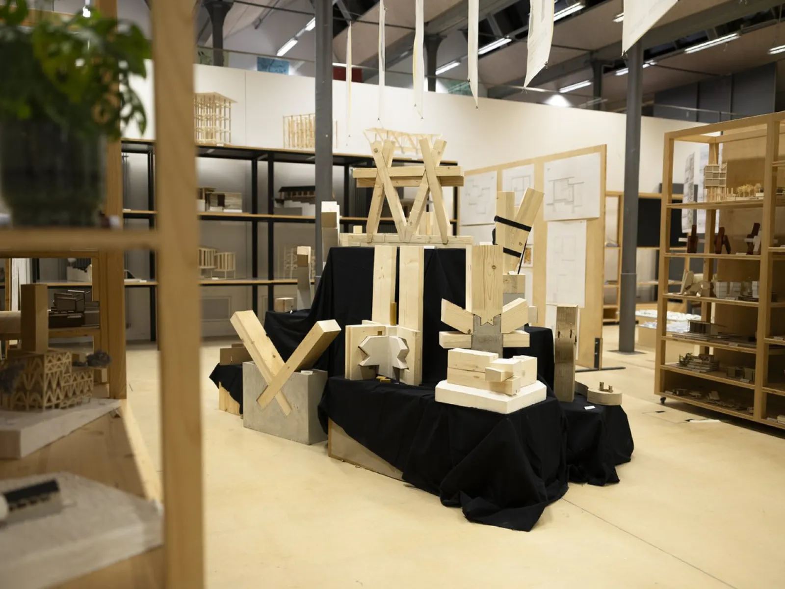Ausstellung von Holzmodellen und Bauteilen in einem modernen Raum. Die Modelle sind auf schwarzen Stoffen arrangiert und zeigen verschiedene architektonische Strukturen. Im Hintergrund sind Regale mit weiteren Modellen und Zeichnungen sichtbar.