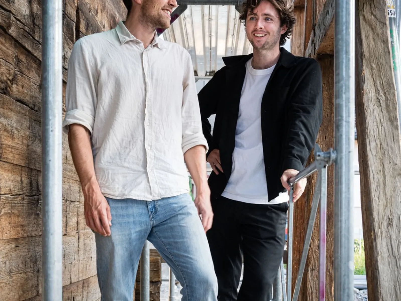 Zwei junge Männer stehen lächelnd auf einem Gerüst in einem Holzbau. Sie tragen legere Kleidung und wirken entspannt. Die Umgebung deutet auf eine Baustelle oder ein architektonisches Projekt hin.