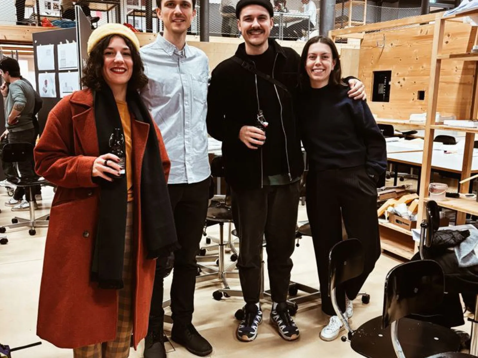 Vier Personen stehen lächelnd in einem Architekturstudio. Sie tragen lässige Kleidung und halten Getränke in der Hand. Im Hintergrund sind Arbeitsplätze und Modelle zu sehen. Die Atmosphäre wirkt freundlich und kreativ.