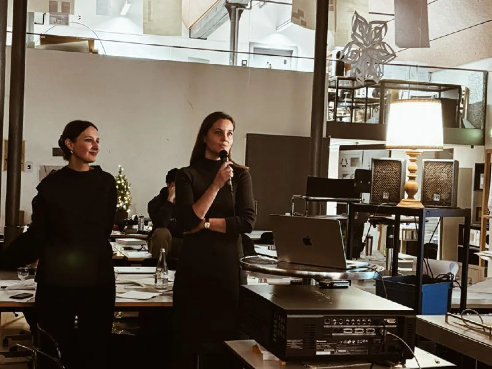 Zwei Personen stehen in einem modernen Architekturstudio. Eine Person hält ein Mikrofon und spricht, während die andere zuhört. Im Hintergrund sind Arbeitsplätze und Architekturmodelle zu sehen.