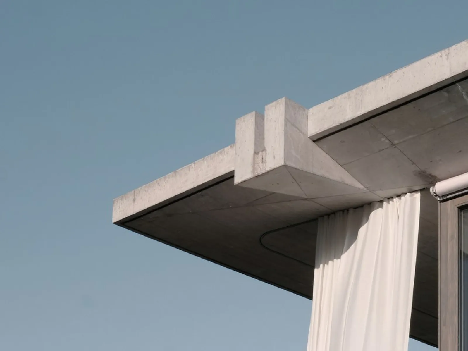 Betonvordach mit klaren Linien und hängendem weißen Vorhang vor blauem Himmel. Moderne Architektur, die Minimalismus und Eleganz vereint.