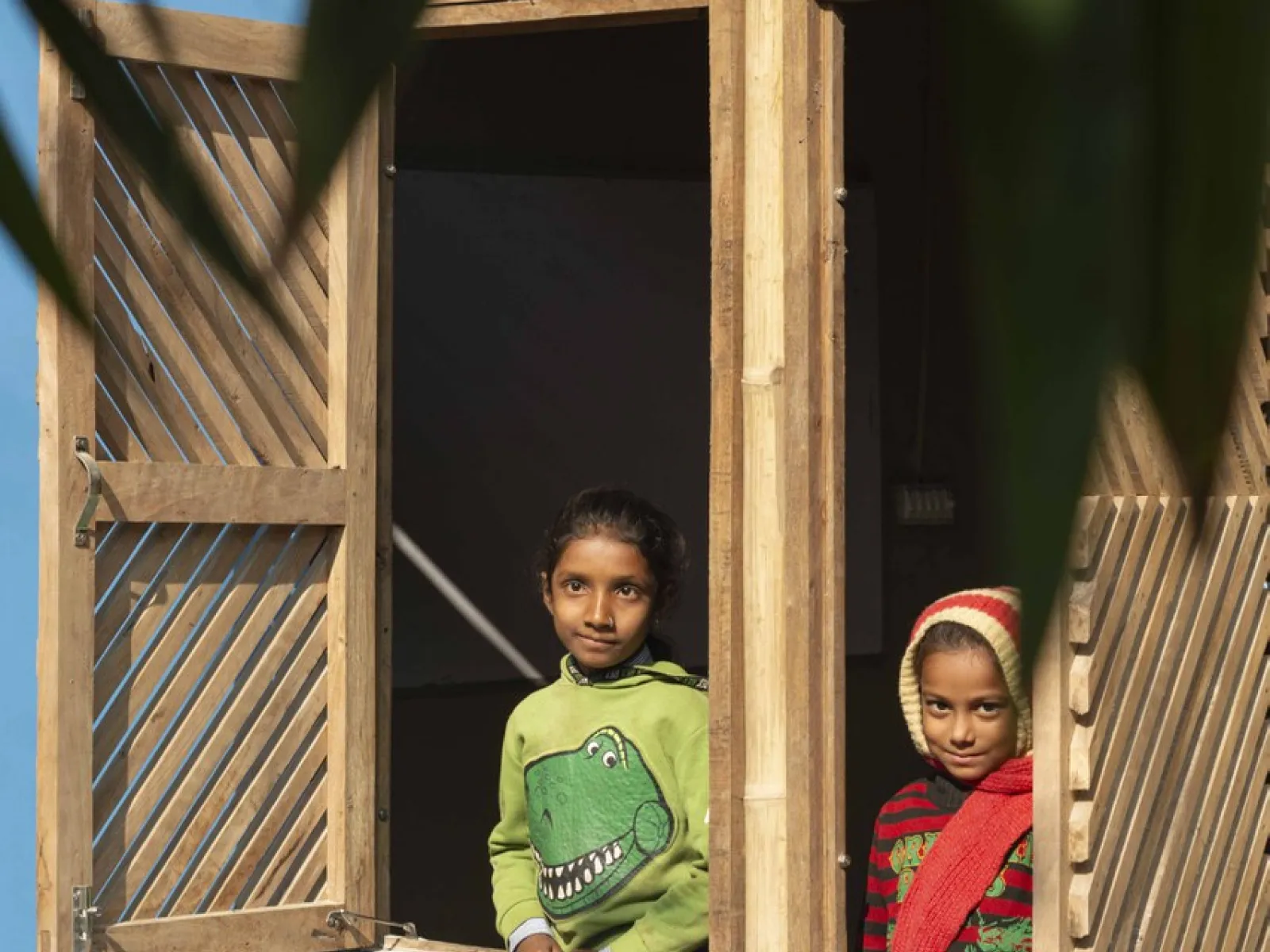 Zwei Kinder schauen aus einem offenen Fenster eines Gebäudes mit blauer Wand und dekorativem Muster. Das Fenster hat hölzerne Fensterläden. Im Vordergrund sind unscharfe Blätter zu sehen.
