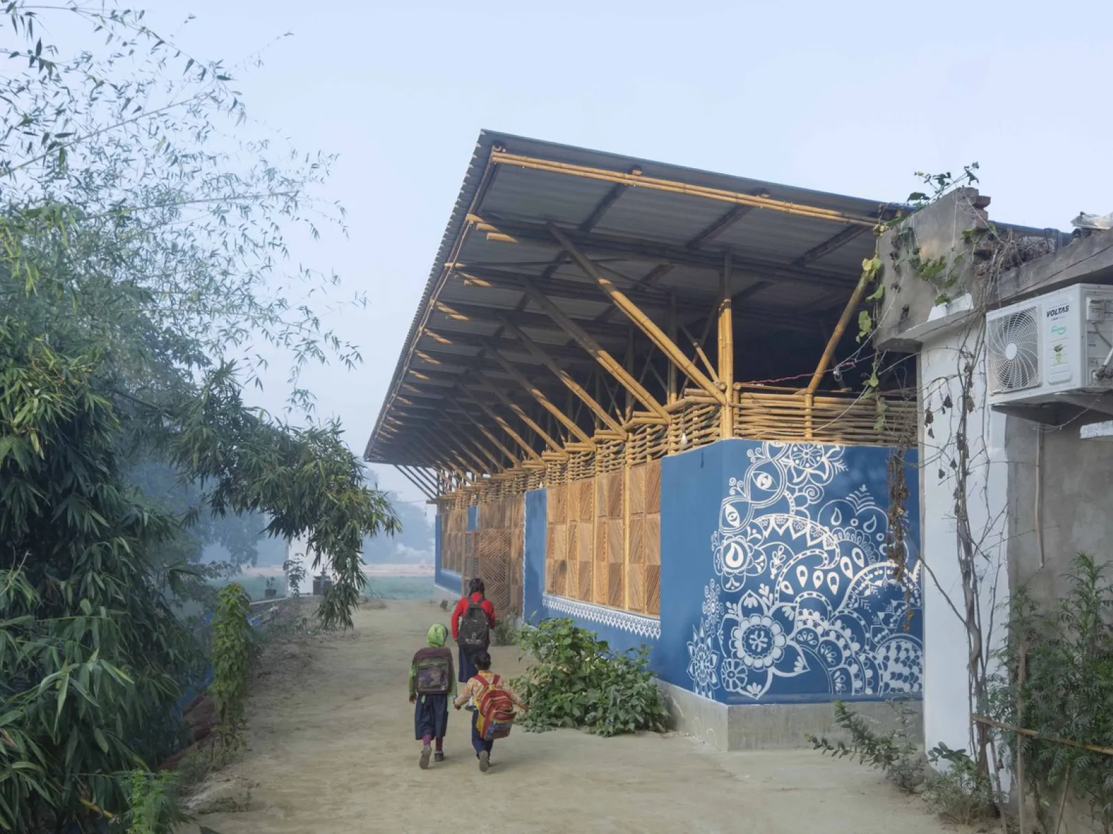 Ein modernes Gebäude mit einem Dach aus Bambus und Wellblech, verziert mit blauen, traditionellen Mustern. Im Vordergrund gehen mehrere Personen auf einem sandigen Weg entlang, umgeben von grüner Vegetation.