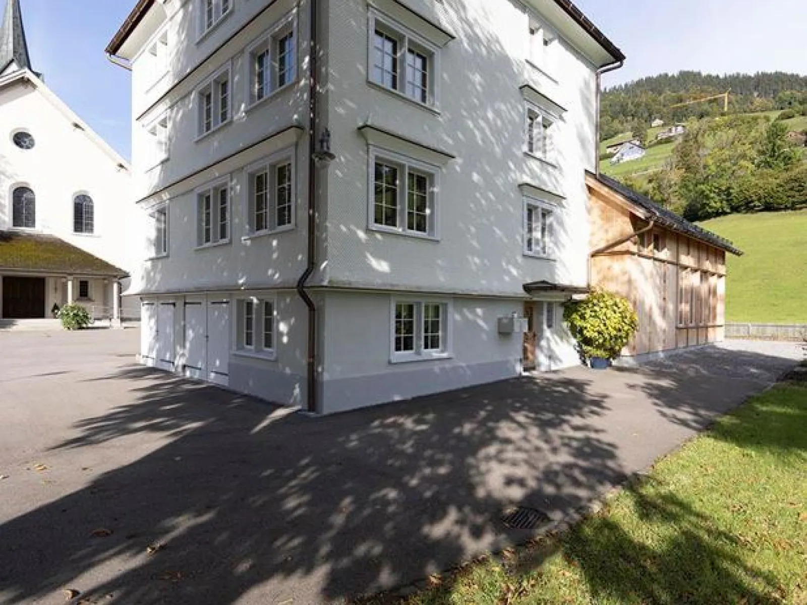 Ein weißes, mehrstöckiges Gebäude mit traditionellen Fenstern steht neben einer Kirche im ländlichen St. Galler Rheintal. Im Hintergrund sind grüne Hügel zu sehen. Der Schatten eines Baumes fällt auf den gepflasterten Vorplatz.