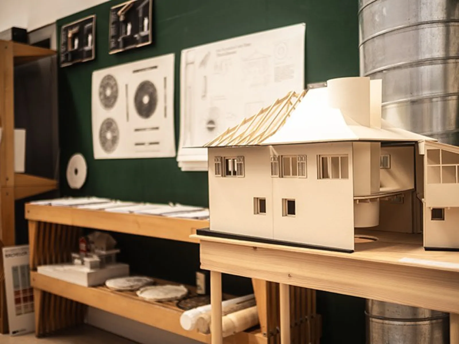 Architekturmodell eines Hauses auf einem Tisch in einem Studio der Universität Liechtenstein. Im Hintergrund sind Architekturpläne und Zeichnungen an einer grünen Wand zu sehen.