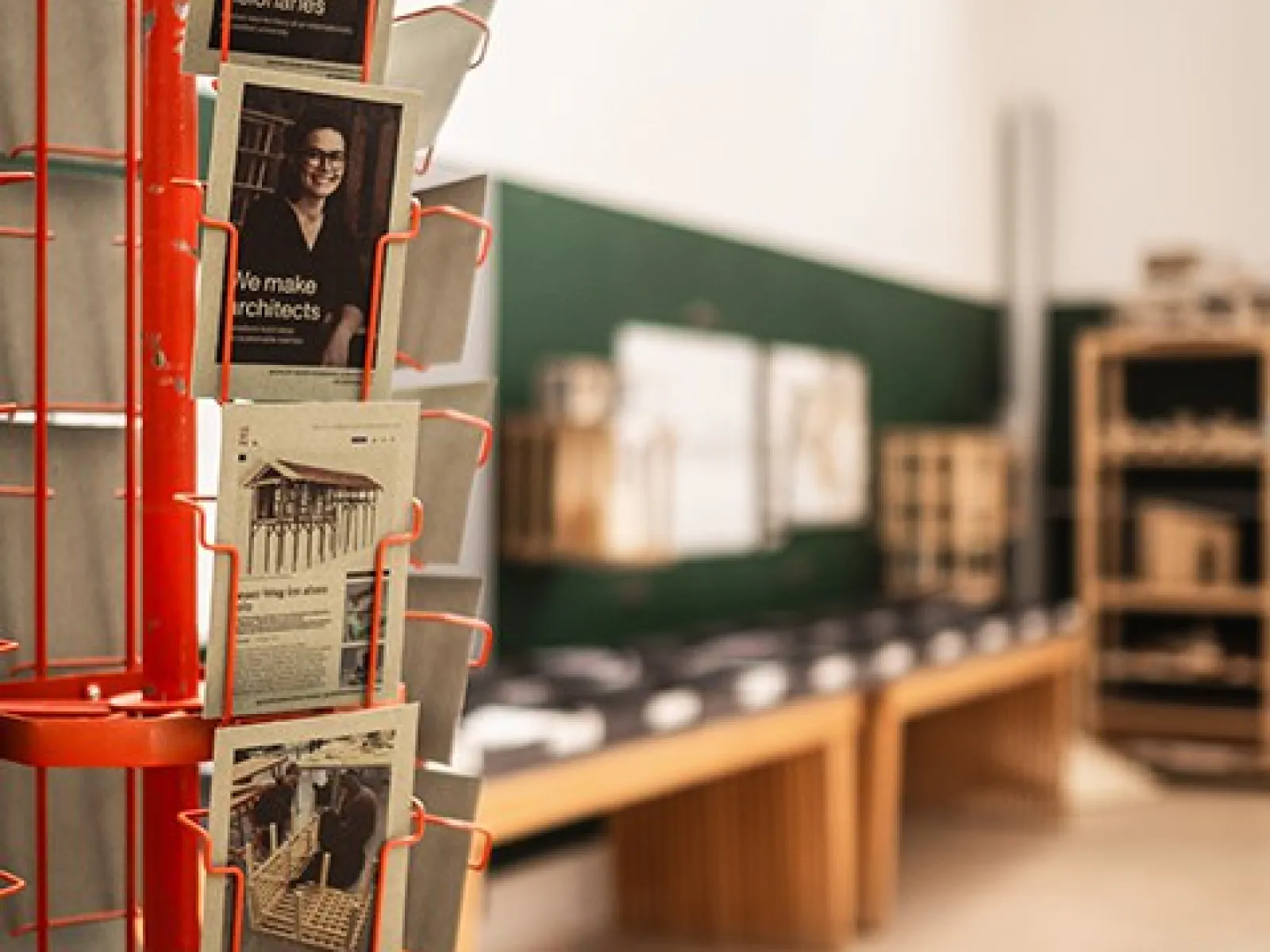 Ein roter Postkartenständer mit Architekturpostkarten im Vordergrund, im Hintergrund ein Raum mit Architekturmodellen und Zeichnungen auf einem Tisch.