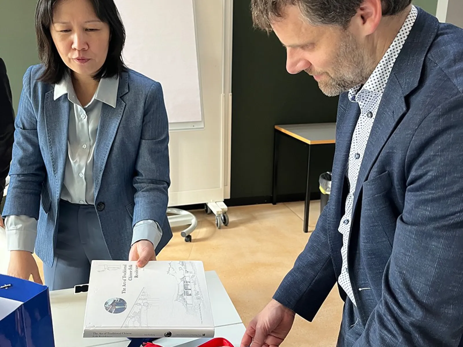 Zwei Personen in formeller Kleidung betrachten Bücher auf einem Tisch in einem Konferenzraum. Im Hintergrund ist ein Flipchart zu sehen.
