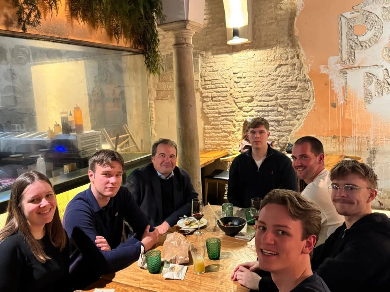 Eine Gruppe von sieben Personen sitzt an einem Tisch in einem Restaurant. Sie lächeln in die Kamera. Im Hintergrund sind eine gemauerte Wand und Pflanzen zu sehen.