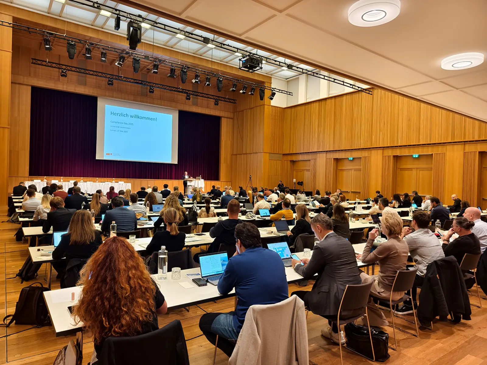 Ein großer Konferenzraum mit vielen Teilnehmern, die an Tischen sitzen und einem Vortrag folgen. Eine Präsentation wird auf einer Leinwand gezeigt. Die Veranstaltung findet in einem modernen Saal mit Holzwänden statt.