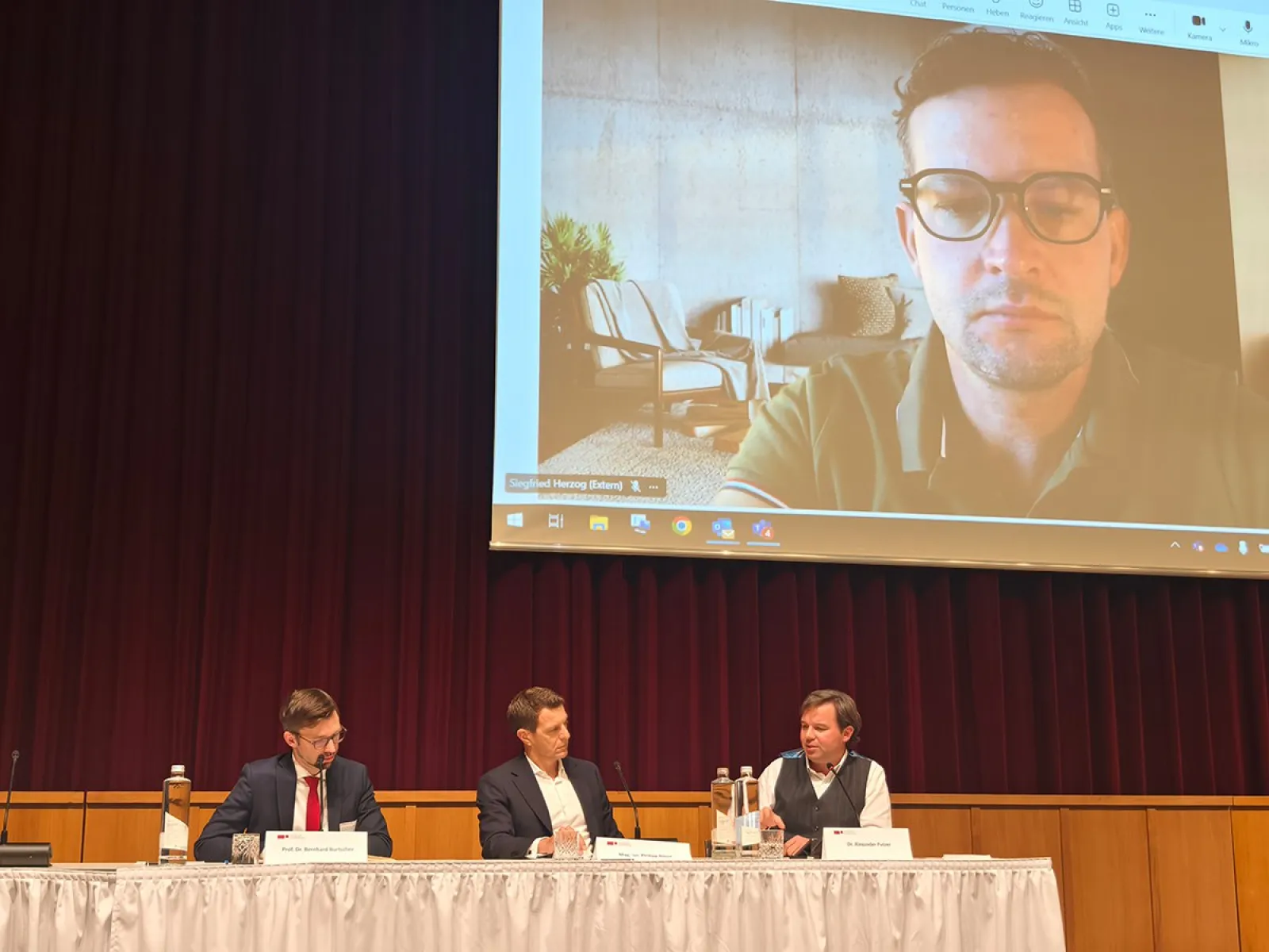 Ein Podium mit drei Männern, die an einem Tisch sitzen und diskutieren. Im Hintergrund ist ein großer Bildschirm mit dem Bild eines weiteren Mannes in einer Videokonferenz zu sehen. Die Szene findet in einem Konferenzsaal statt.