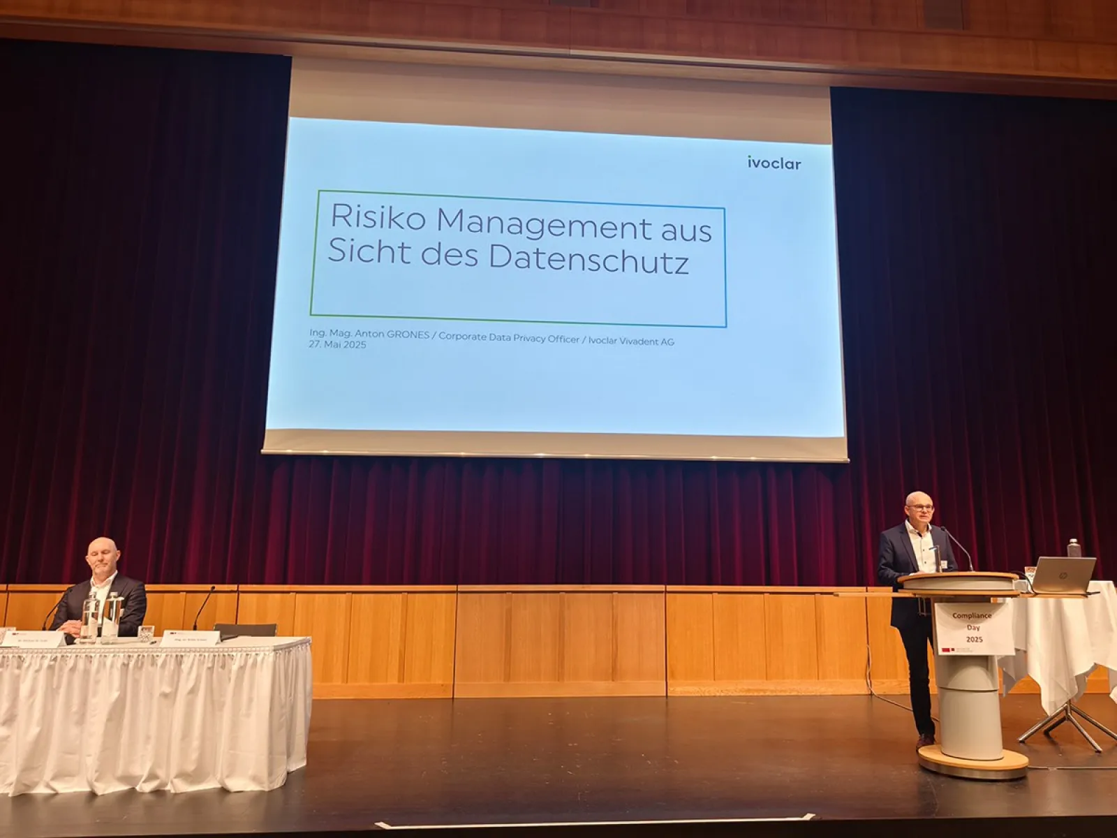 Ein Redner steht an einem Podium auf einer Bühne und hält eine Präsentation mit dem Titel "Risiko Management aus Sicht des Datenschutzes". Ein weiterer Teilnehmer sitzt links am Tisch. Der Hintergrund ist ein roter Vorhang.