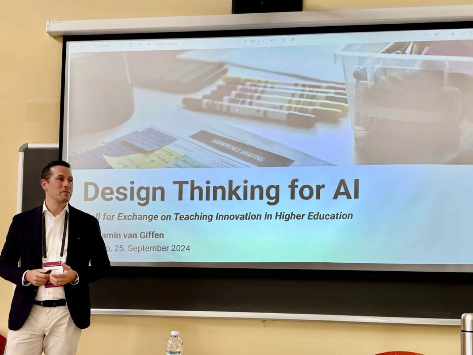 Ein Mann steht vor einer Leinwand, auf der eine Präsentation mit dem Titel "Design Thinking for AI" gezeigt wird. Der Raum wirkt wie ein Seminarraum, und es sind einige Stifte und Notizen auf der Leinwand zu sehen.