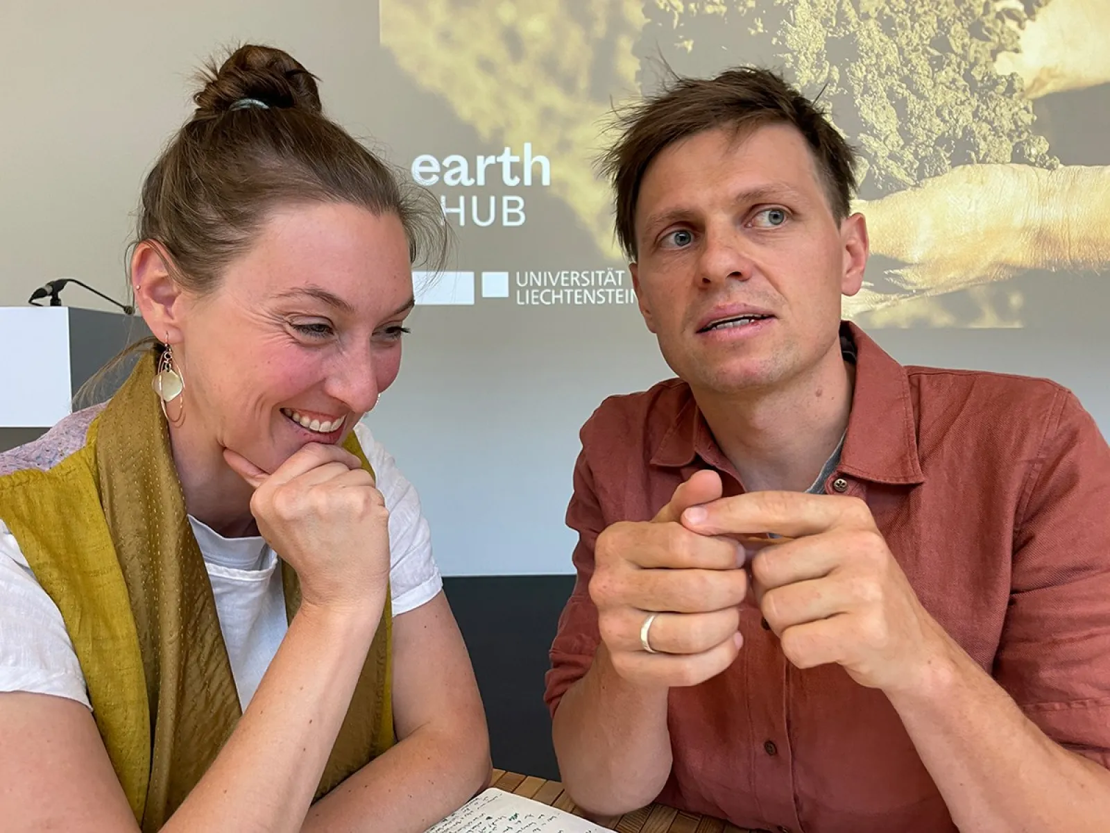 Zwei Personen sitzen an einem Tisch und diskutieren. Im Hintergrund ist ein Bildschirm mit dem Logo des Earth-Hub-Symposiums der Universität Liechtenstein zu sehen.