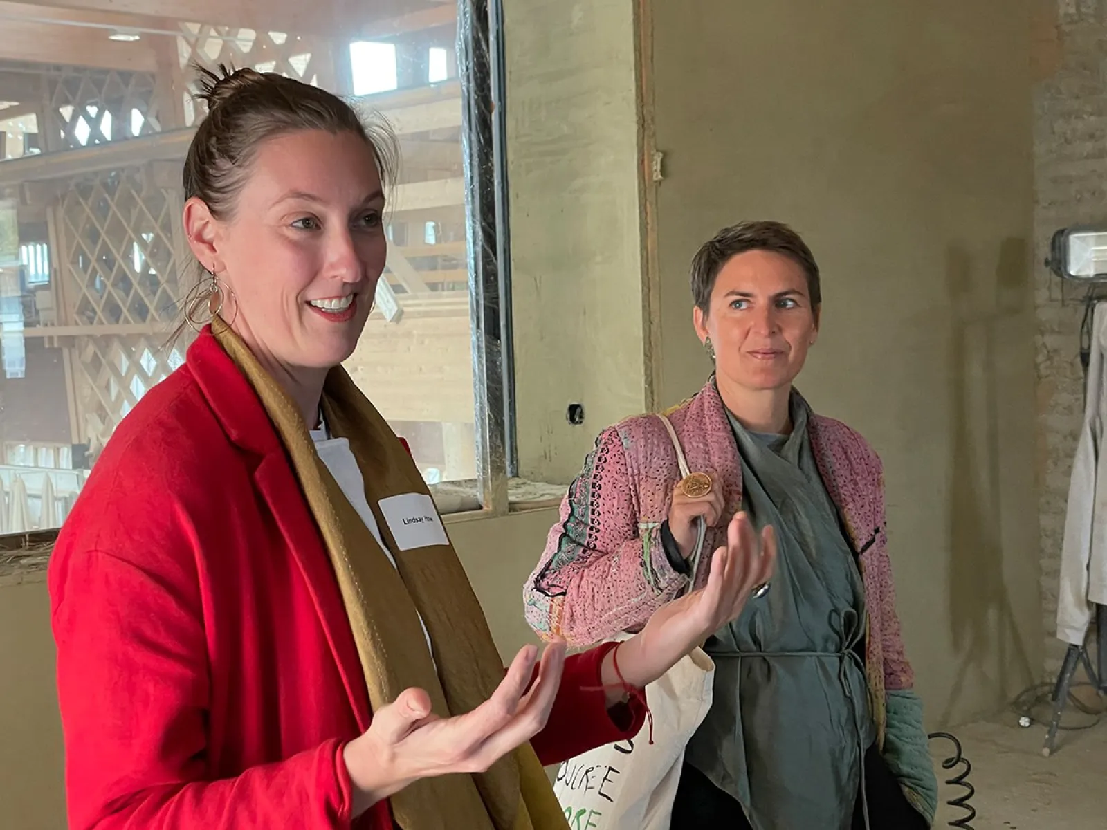 Zwei Personen stehen in einem Raum mit Lehmwänden. Eine Person trägt einen roten Blazer und gestikuliert, während die andere Person aufmerksam zuhört. Im Hintergrund sind Holzelemente sichtbar.