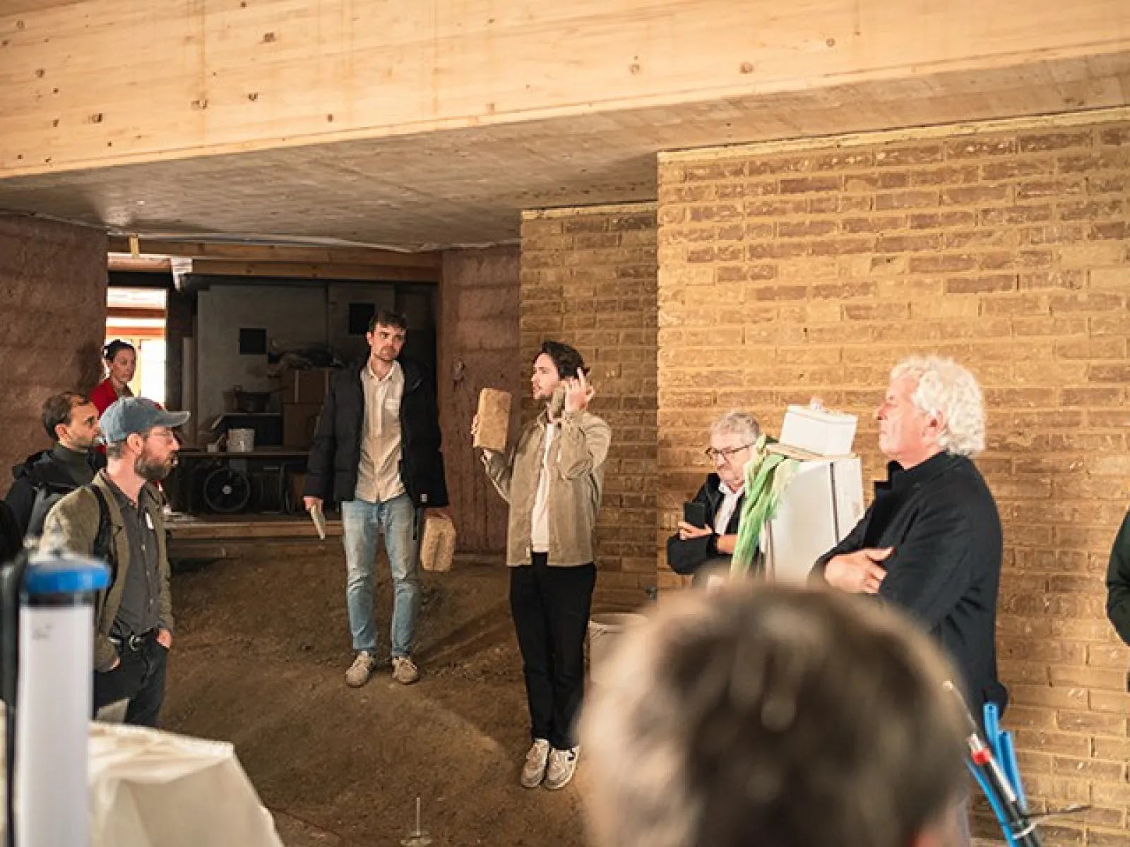 Eine Gruppe von Personen steht in einem Raum mit Wänden aus Stampflehm und Holz. Eine Person hält ein Präsentationsobjekt und spricht zu den anderen. Die Szene zeigt eine Diskussion über nachhaltige Baumaterialien bei einem Symposium der Liechtenstein School of Architecture.