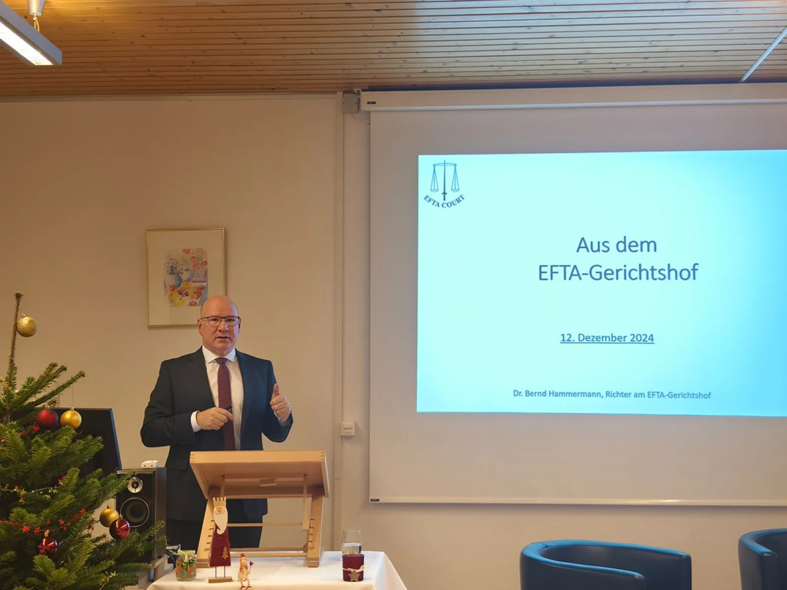 Ein Redner steht neben einem Pult und präsentiert vor einer Leinwand mit der Aufschrift „Aus dem EFTA-Gerichtshof“ bei einer Veranstaltung an der Universität Liechtenstein. Links im Bild ist ein geschmückter Weihnachtsbaum zu sehen.