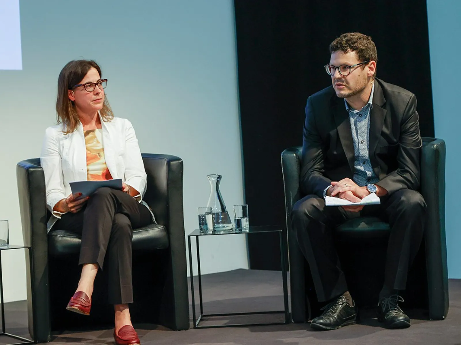 Zwei Personen sitzen auf einer Bühne in schwarzen Sesseln. Die Frau links trägt eine weiße Jacke und hält Papiere, der Mann rechts trägt einen dunklen Anzug. Zwischen ihnen steht ein Wasserglas auf einem kleinen Tisch.