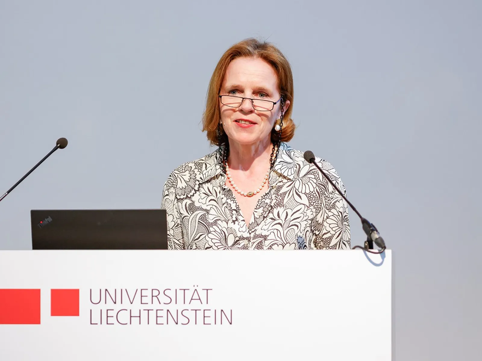 Es ist eine Frau zu sehen, welche vor einem Redepult mit dem Logo der Universität Liechtenstein steht.