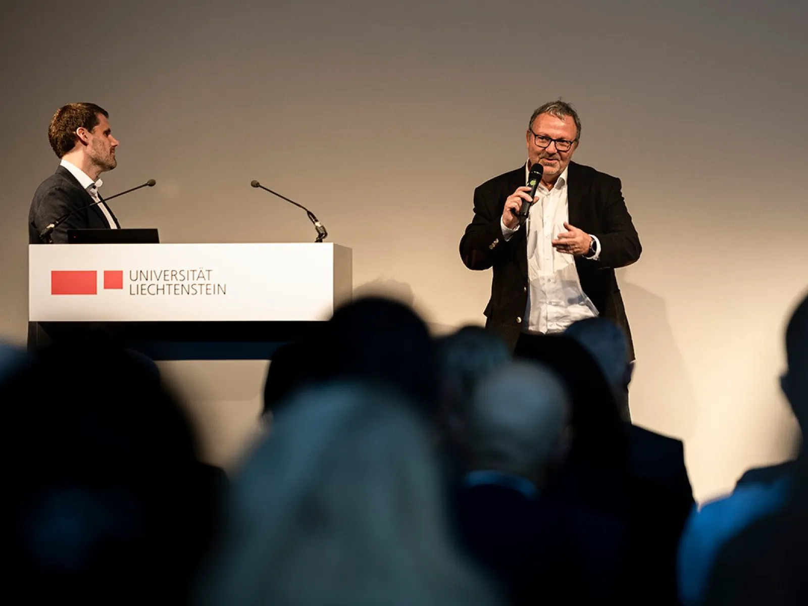 Zwei Männer stehen auf einer Bühne bei einer Veranstaltung an der Universität Liechtenstein. Einer spricht in ein Mikrofon, während der andere neben einem Rednerpult mit dem Logo der Universität steht. Im Vordergrund sind unscharf die Köpfe des Publikums zu sehen.