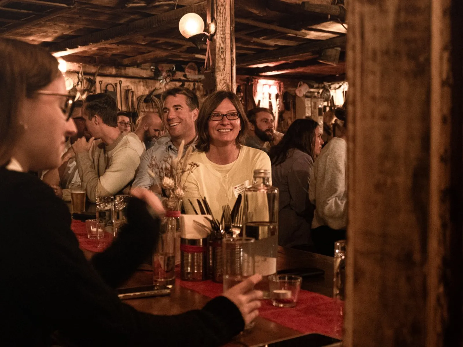 Eine Gruppe von Menschen sitzt in einer gemütlichen, rustikalen Bar. Im Vordergrund unterhalten sich Personen an einem Tisch, während im Hintergrund weitere Gäste lächeln und die Atmosphäre genießen. Die Szene wirkt lebhaft und einladend.