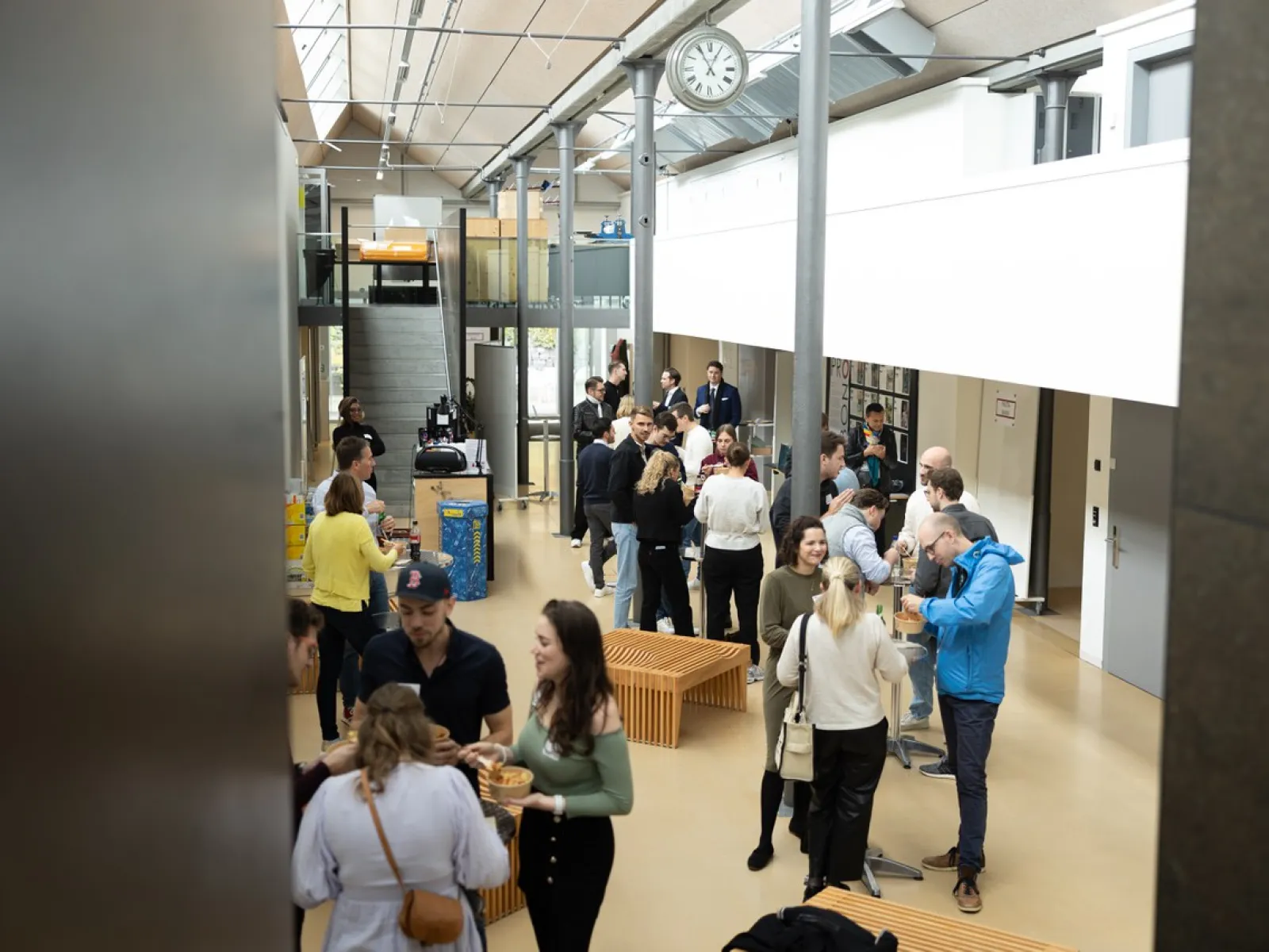 Eine Gruppe von Menschen versammelt sich in einem modernen, lichtdurchfluteten Innenraum der Universität Liechtenstein. Sie stehen in kleinen Gruppen zusammen und unterhalten sich. Im Hintergrund sind Treppen und eine Uhr zu sehen.