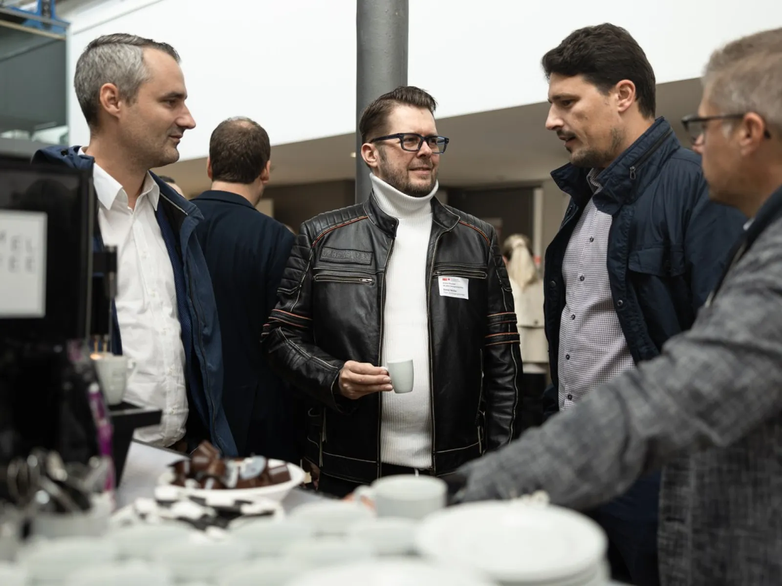 Eine Gruppe von vier Männern steht an einem Stehtisch und unterhält sich bei einer Veranstaltung. Einer hält eine Tasse Kaffee. Im Vordergrund sind Geschirr und Snacks zu sehen. Die Atmosphäre wirkt gesellig und professionell.