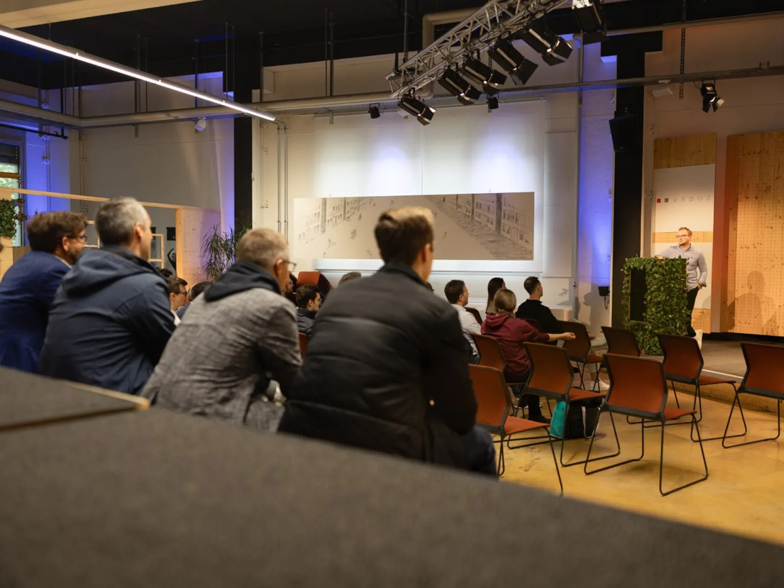 Eine Gruppe von Menschen sitzt in einem Veranstaltungsraum und hört einem Vortrag zu. Im Hintergrund ist ein Redner am Podium zu sehen. Der Raum ist modern gestaltet und gut beleuchtet.