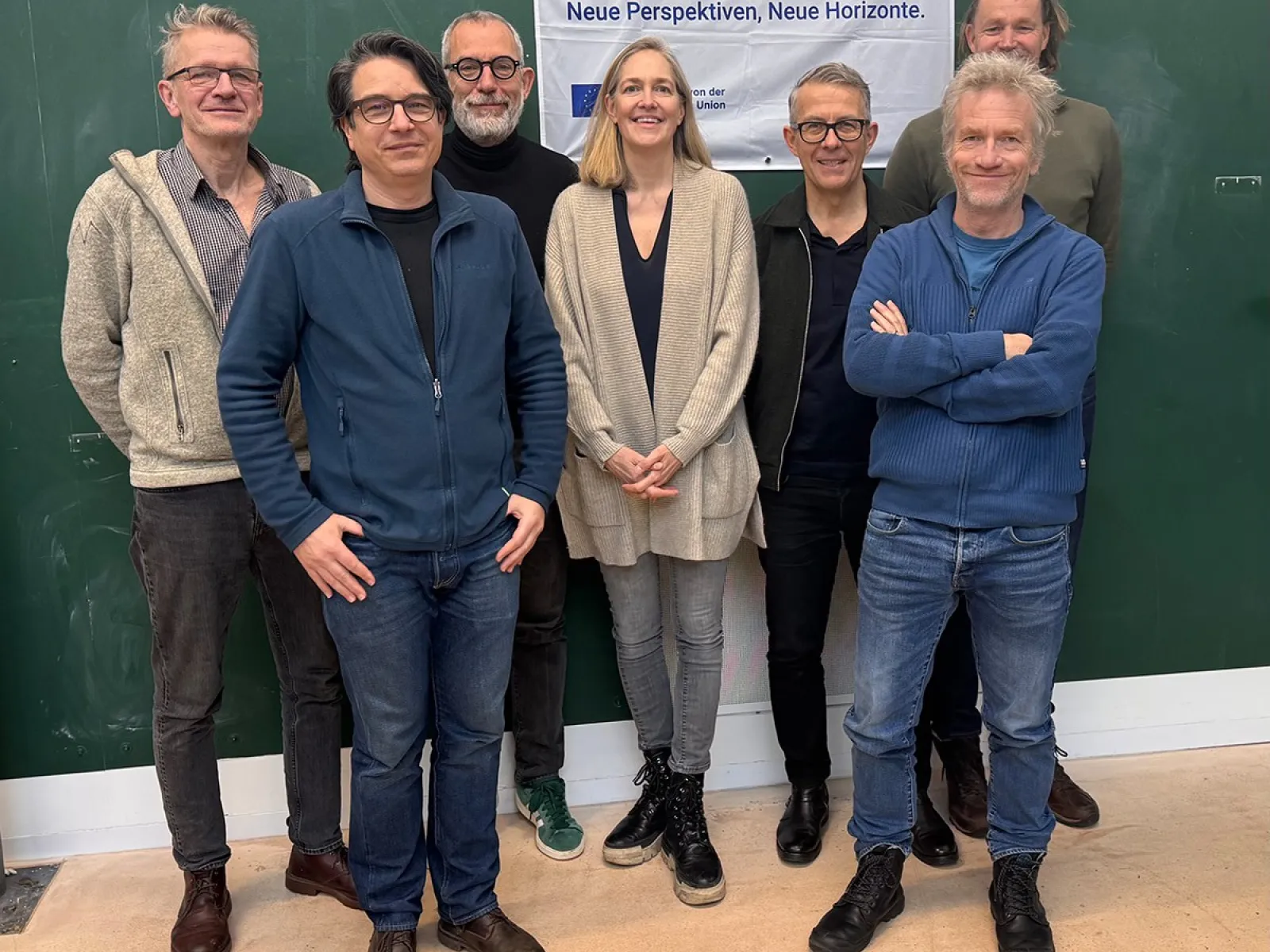 Eine Gruppe von acht Personen steht vor einem grünen Hintergrund. Sie lächeln in die Kamera. Im Hintergrund hängt ein Banner mit der Aufschrift "Erasmus+ Neue Perspektiven, Neue Horizonte".