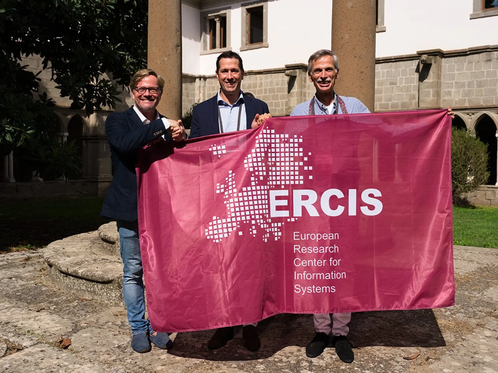 Drei Personen halten ein Banner mit der Aufschrift "ERCIS - European Research Center for Information Systems" in einem sonnigen Innenhof. Im Hintergrund sind historische Gebäude und Säulen zu sehen.