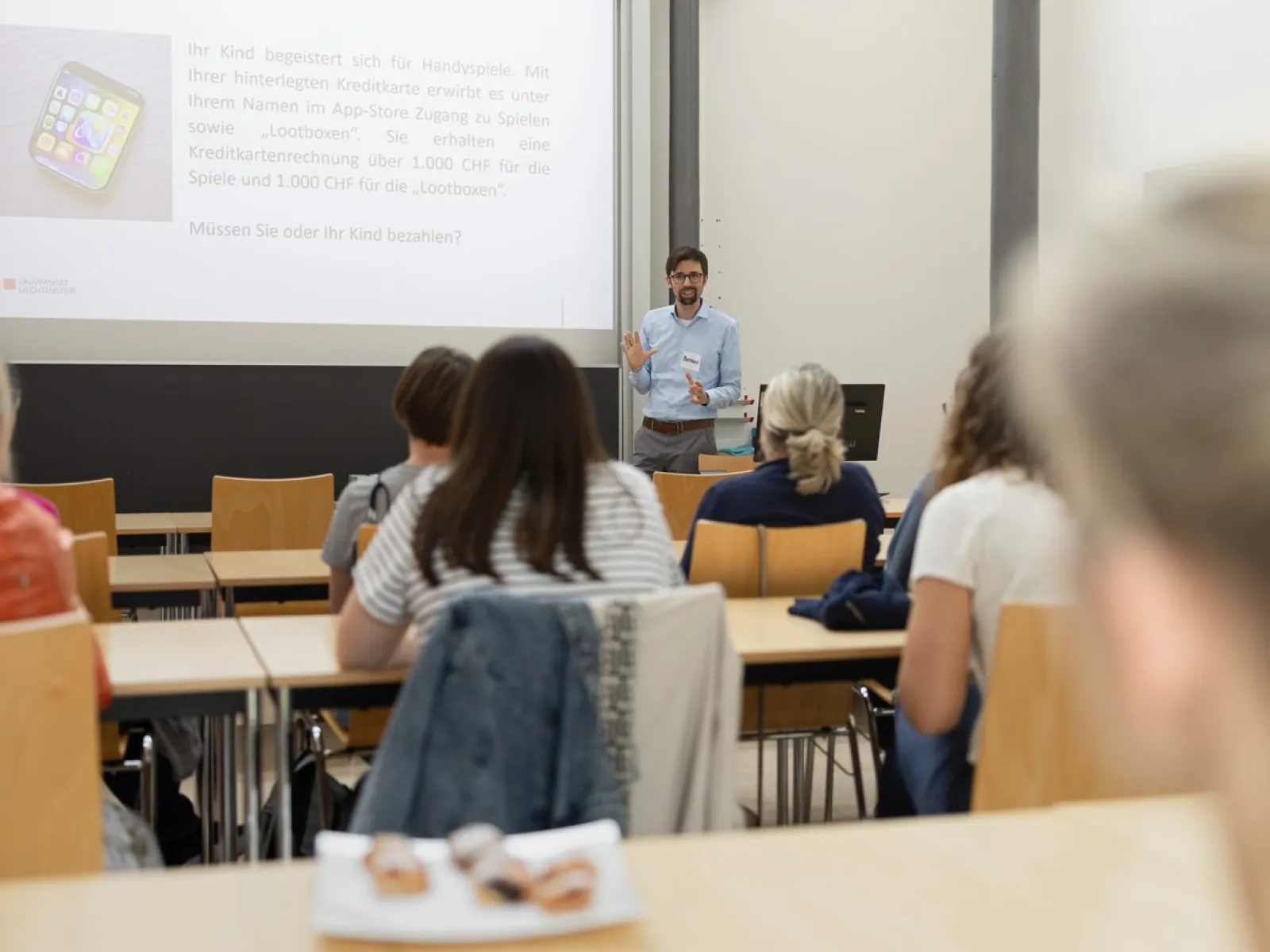 Ein Dozent hält einen Vortrag in einem Seminarraum vor mehreren Erwachsenen. Die Zuhörer sitzen an Tischen und schauen zur Präsentation an der Leinwand.