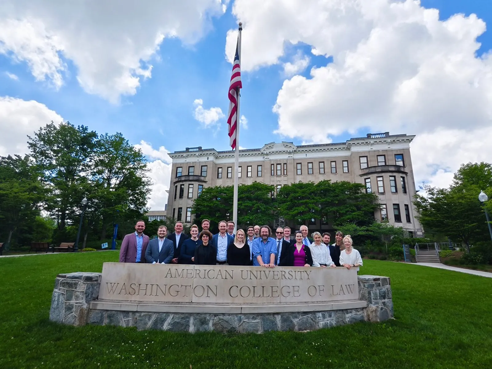 Gruppe von Studierenden und Dozenten des Executive Master of Laws der Universität Liechtenstein vor dem Washington College of Law der American University, mit einer US-Flagge im Vordergrund und einem historischen Gebäude im Hintergrund.