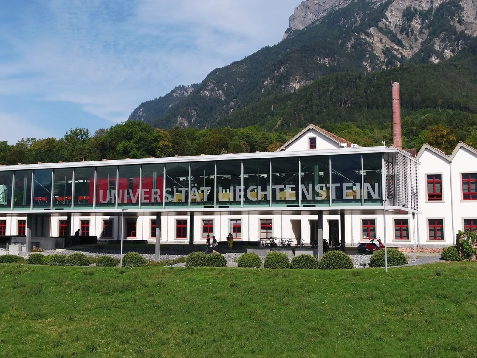 Außenansicht der Universität Liechtenstein mit modernem Glasgebäude und traditionellem weißen Gebäude im Hintergrund, umgeben von grüner Landschaft und Bergen.