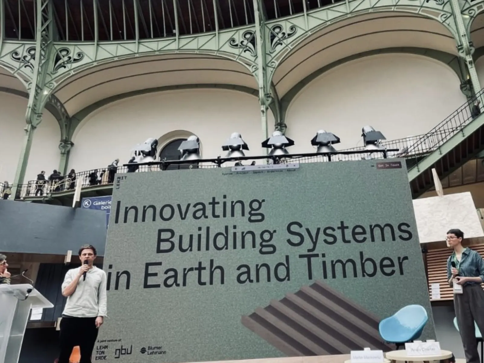 Ein Redner präsentiert auf einer Bühne vor einem großen Bildschirm mit der Aufschrift „Innovating Building Systems in Earth and Timber“ im Grand Palais, Paris. Im Hintergrund ist die beeindruckende Architektur des Veranstaltungsortes zu sehen.