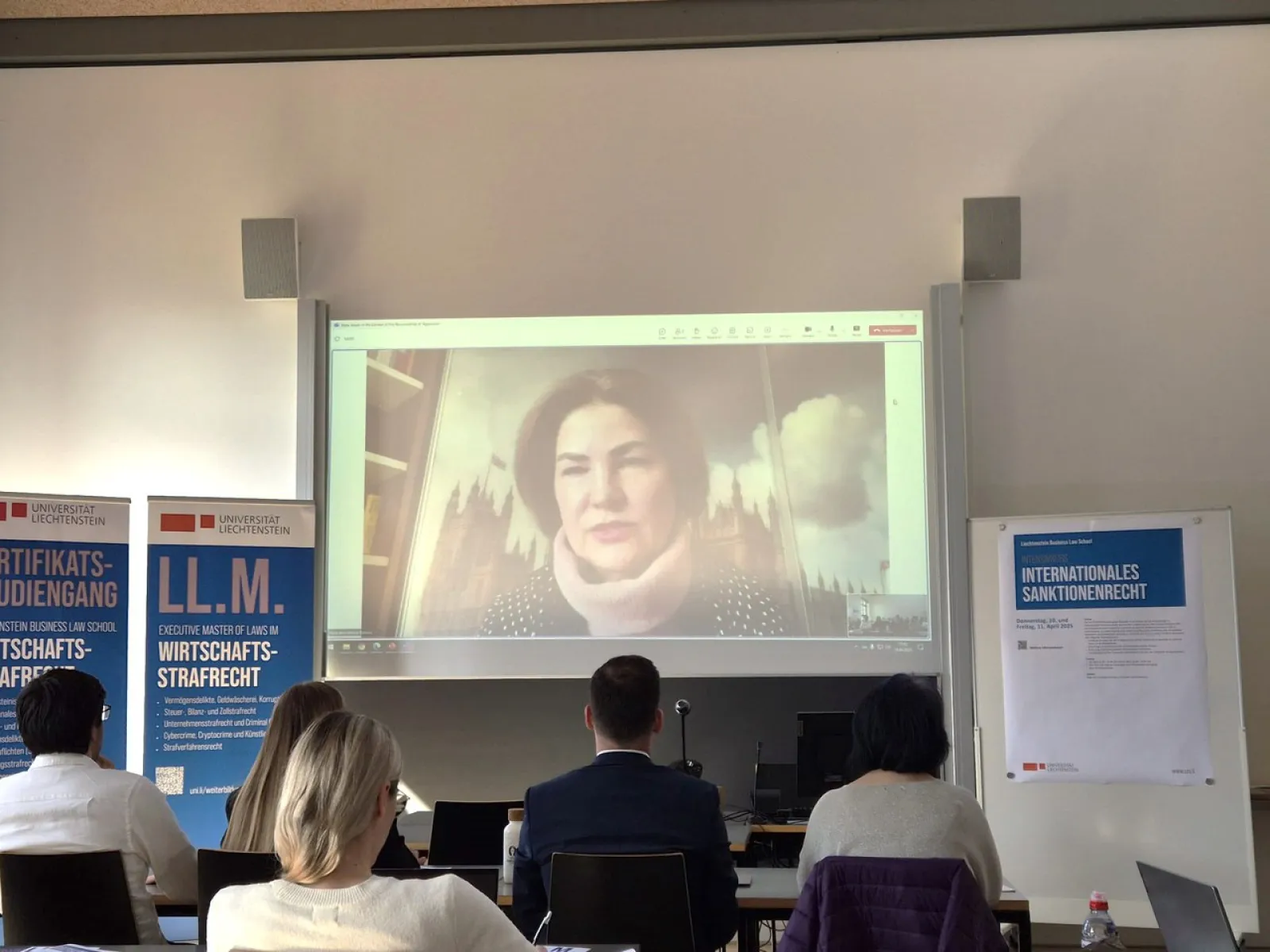Vortrag im Rahmen des Intensivkurses „Internationales Sanktionenrecht“ an der Universität Liechtenstein. Eine Rednerin wird per Videokonferenz auf eine Leinwand projiziert. Im Vordergrund sitzen Teilnehmende, im Hintergrund sind Banner der Universität zu sehen.