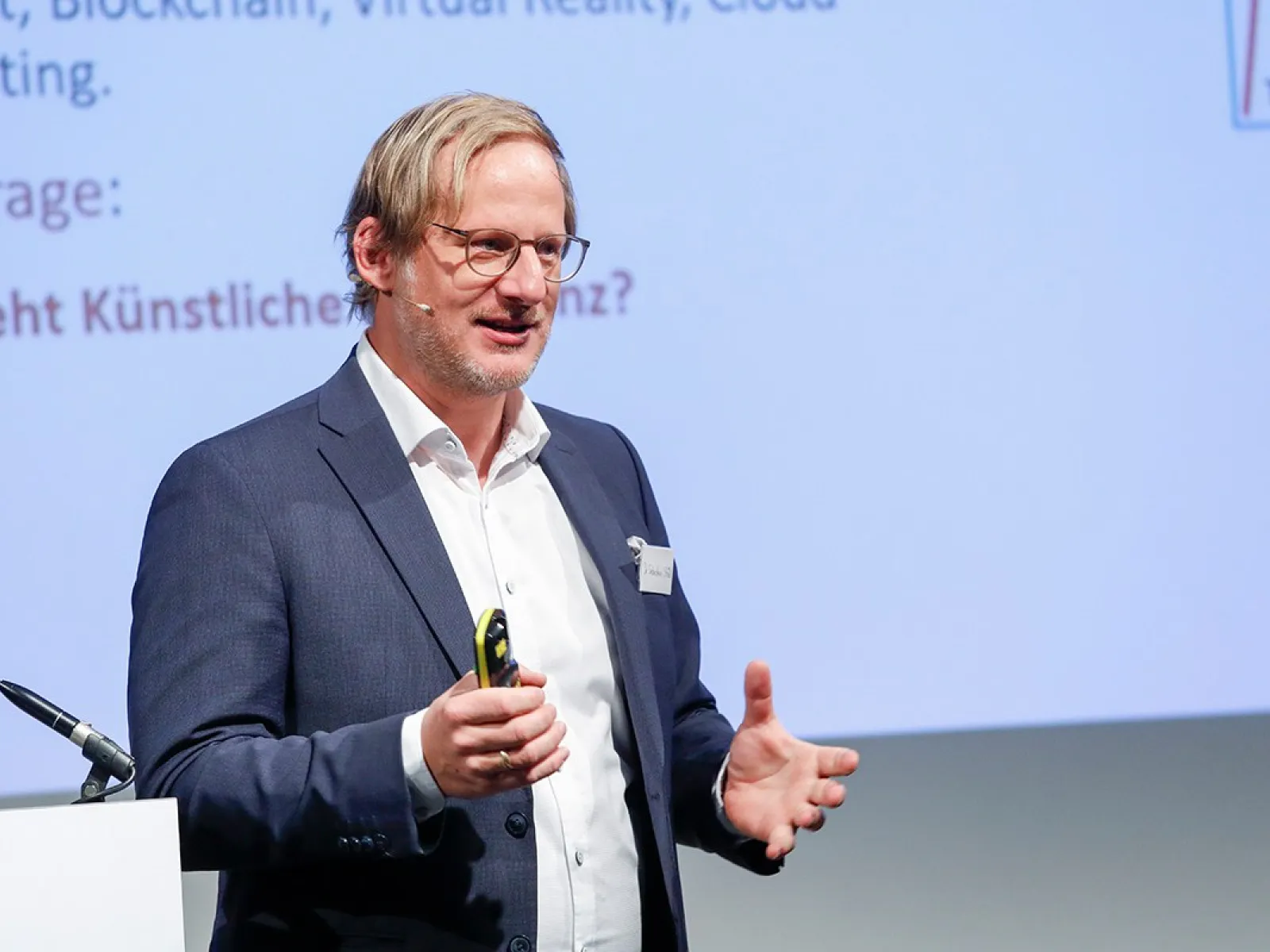 Ein Redner in einem Anzug hält einen Vortrag auf einer Konferenz über Künstliche Intelligenz an der Universität Liechtenstein. Im Hintergrund ist eine Präsentationsfolie zu sehen.