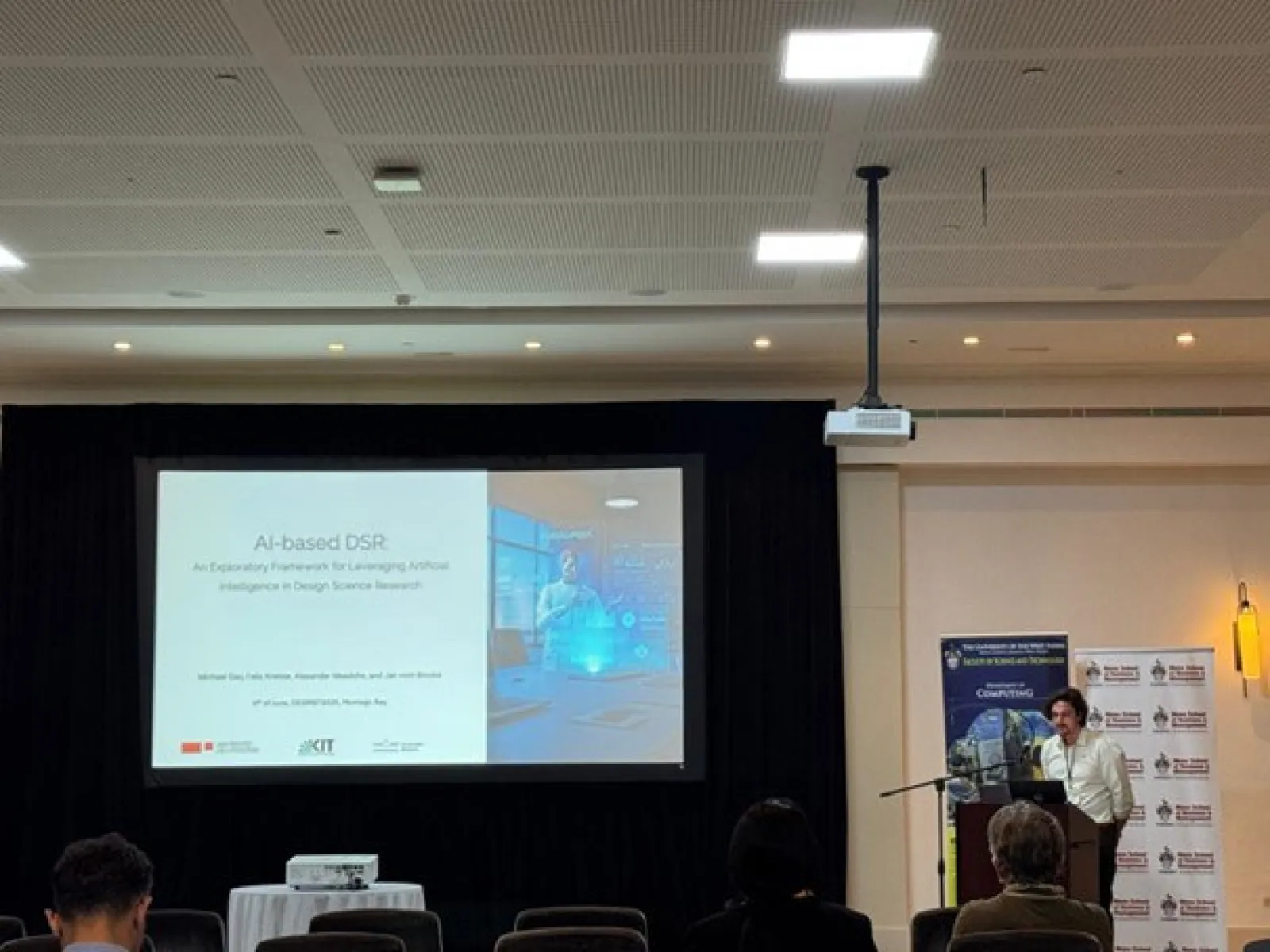 Ein Vortragender präsentiert auf einer Konferenz vor einer Leinwand mit der Folie "AI-based DSR". Im Raum sind mehrere Zuhörer zu sehen.