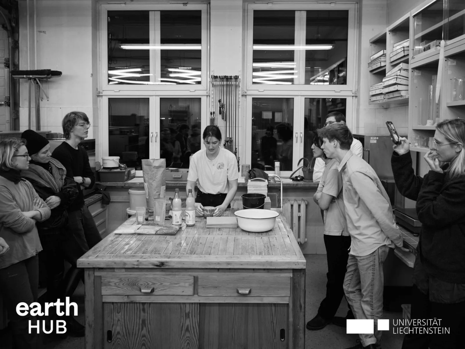 Eine Gruppe von Menschen steht in einem Workshop-Raum um einen Tisch, auf dem Materialien und Werkzeuge liegen. Eine Person in der Mitte erklärt etwas. Im Hintergrund sind Regale und Fenster zu sehen. Logos von "earth HUB" und "Universität Liechtenstein" sind eingeblendet.