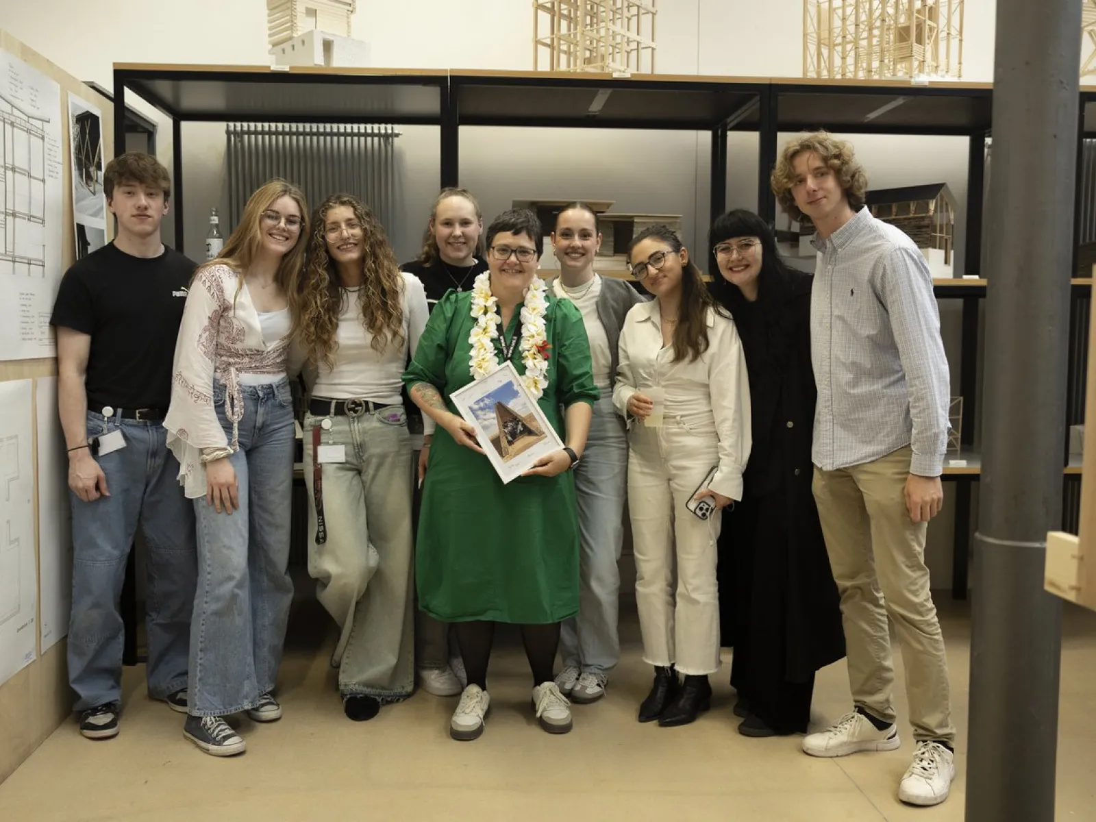 Eine Gruppe von neun Personen steht lächelnd in einem Innenraum. Eine Person in der Mitte hält ein Buch und trägt eine grüne Kleidung mit einer Blumenkette. Im Hintergrund sind architektonische Modelle zu sehen.