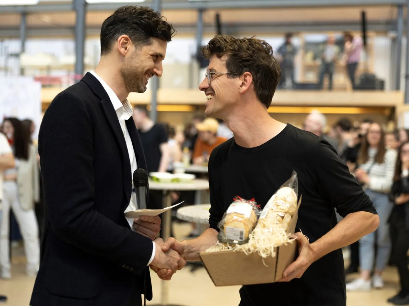 Zwei Männer schütteln sich die Hand bei einer Veranstaltung in einem Innenraum. Einer hält ein Mikrofon, der andere ein Geschenkpaket. Im Hintergrund sind weitere Personen unscharf zu erkennen.