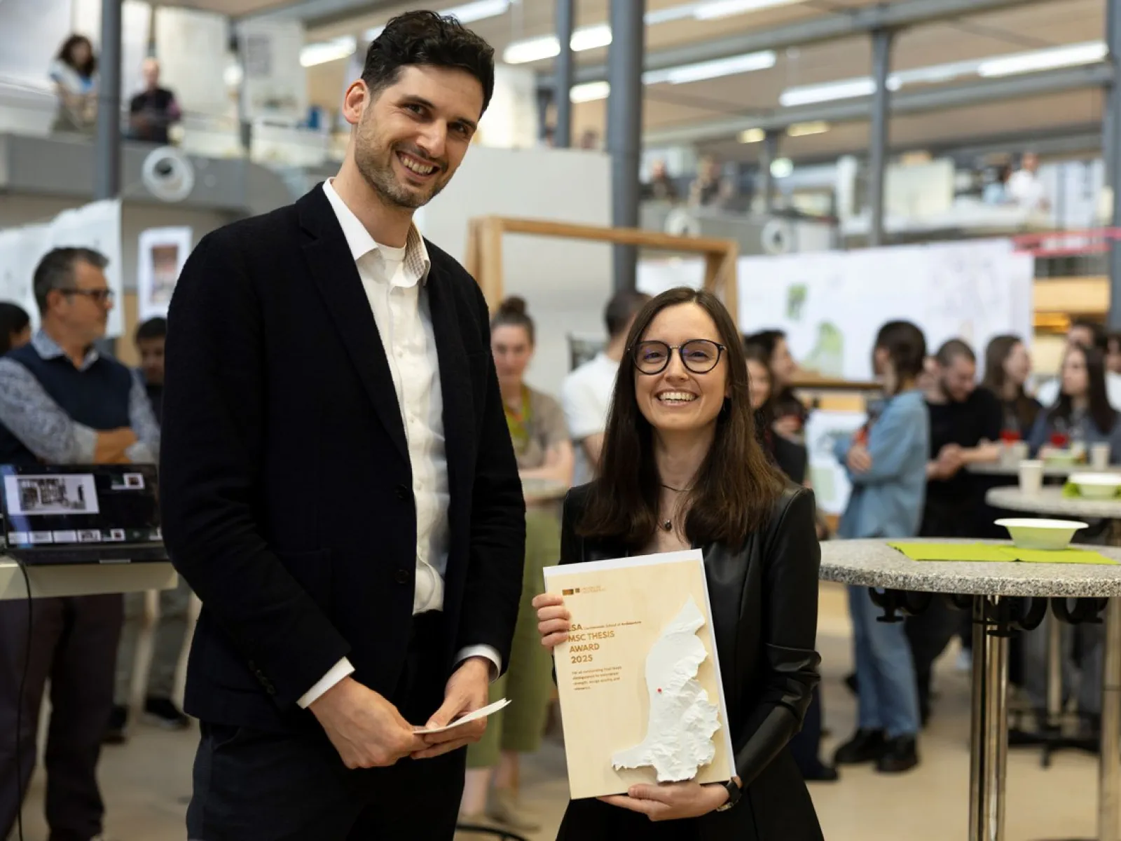 Zwei Personen stehen lächelnd in einem modernen Innenraum während einer Veranstaltung. Eine Person hält ein Buch mit einer Karte auf dem Cover. Im Hintergrund sind weitere Personen und Ausstellungstafeln zu sehen.