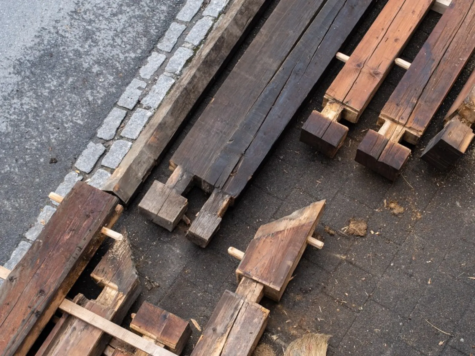 Holzbalken und Bauteile liegen auf einem gepflasterten Boden neben einem Bordstein. Die Materialien sind teilweise bearbeitet und bereit für den Einsatz in einem Bauprojekt. Das Bild zeigt den Fokus auf nachhaltige Baupraktiken und Wiederverwendung von Materialien.