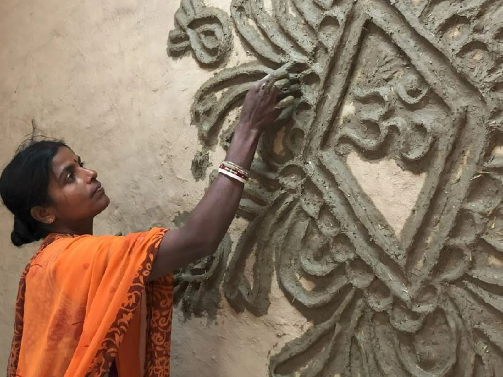 Eine Frau in einem orangefarbenen Sari verziert eine Wand mit traditionellen Tonmustern. Sie arbeitet konzentriert an einem kunstvollen Relief.