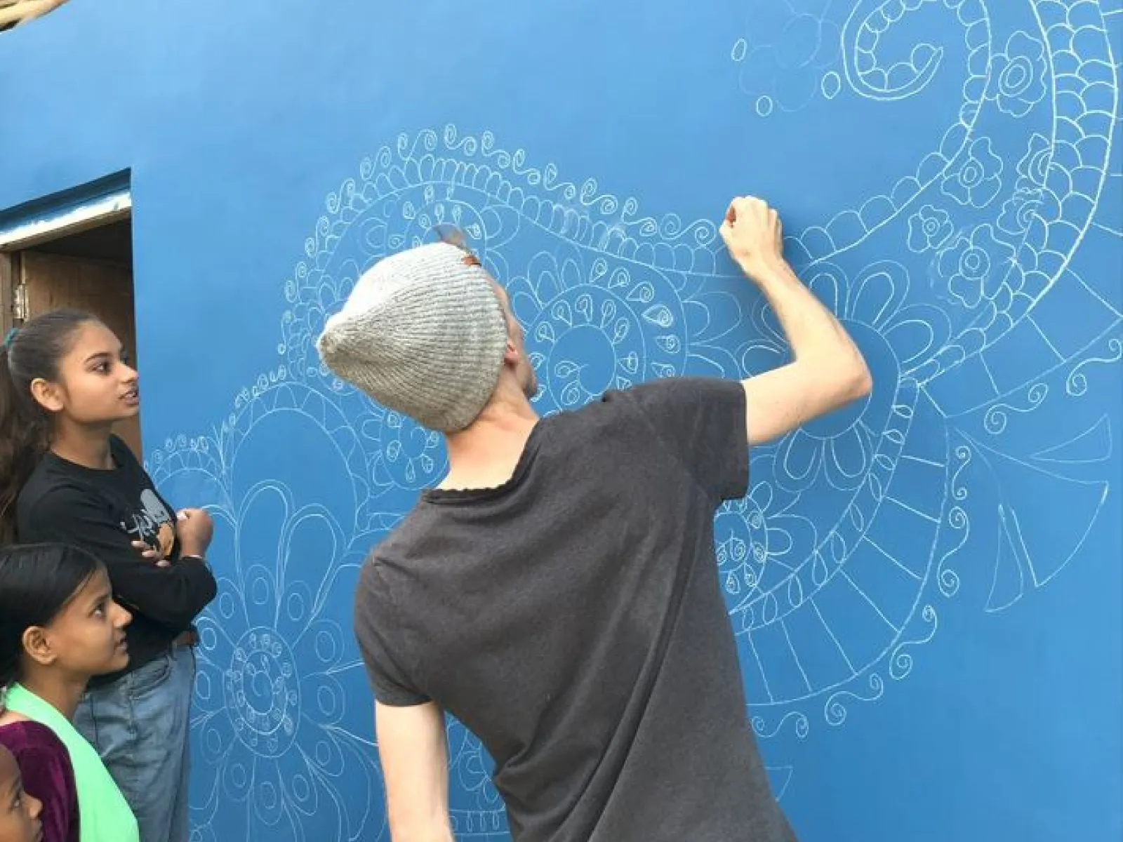 Eine Person zeichnet ein detailliertes Muster auf eine blaue Wand, während drei Kinder zuschauen. Die Szene zeigt gemeinschaftliches Engagement beim Bau einer Schule in Indien.