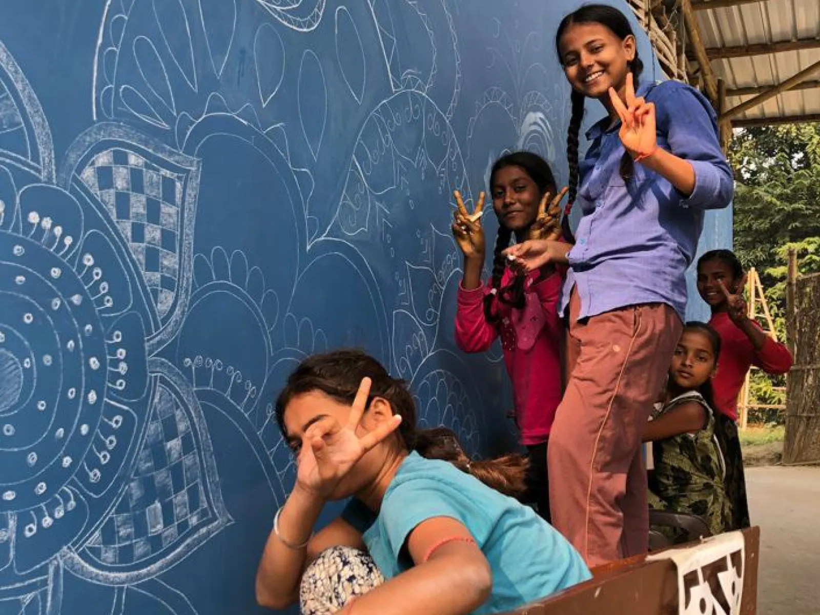 Kinder posieren fröhlich vor einer blau bemalten Wand mit kunstvollen weißen Mustern. Sie zeigen mit den Fingern das Friedenszeichen. Die Szene vermittelt Freude und Gemeinschaftsgefühl.
