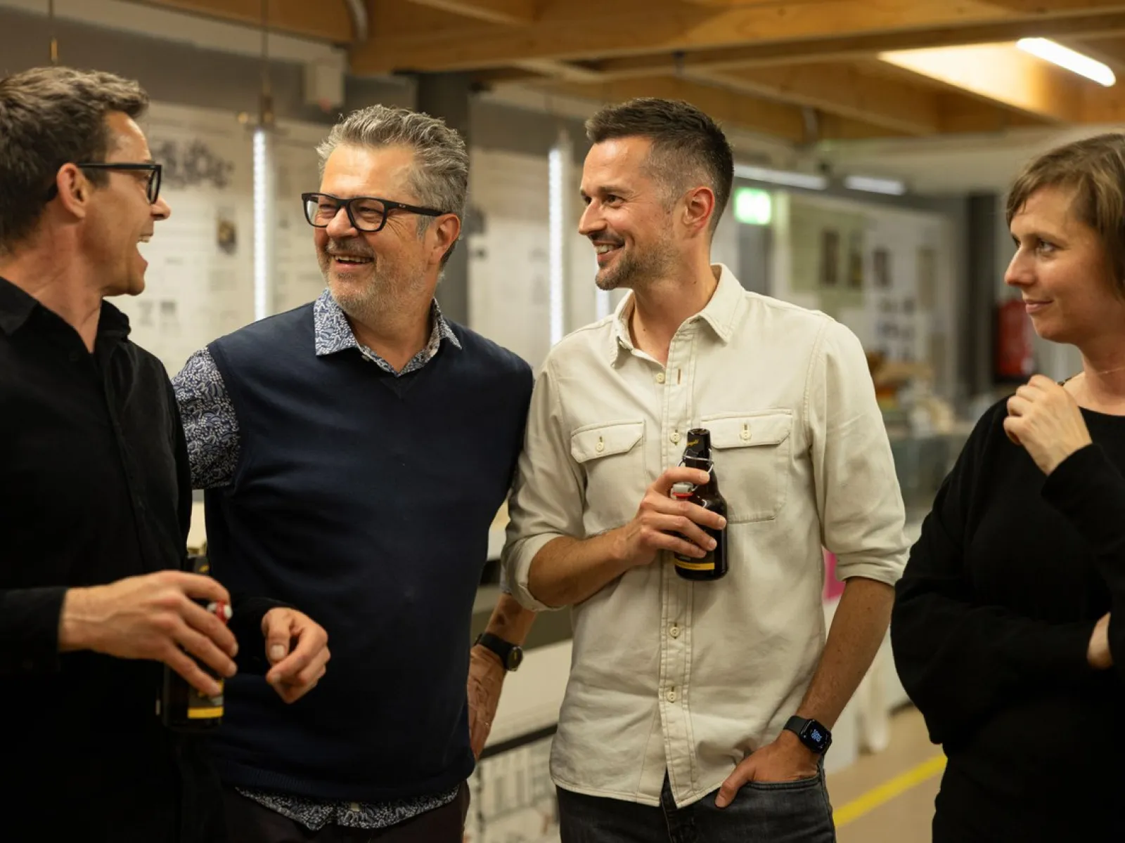 Vier Personen stehen in einem Innenraum und unterhalten sich fröhlich. Zwei von ihnen halten Getränke in der Hand. Im Hintergrund sind Ausstellungstafeln und eine Holzkonstruktion zu sehen. Die Atmosphäre wirkt entspannt und gesellig.